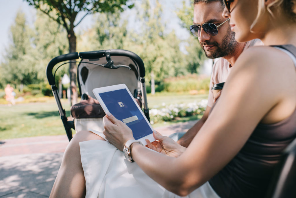 parents sitting near baby carriage in park and looking at tablet with loaded facebook page