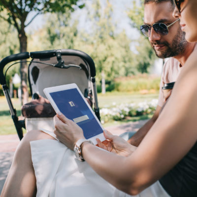 parents sitting near baby carriage in park and looking at tablet with loaded facebook page